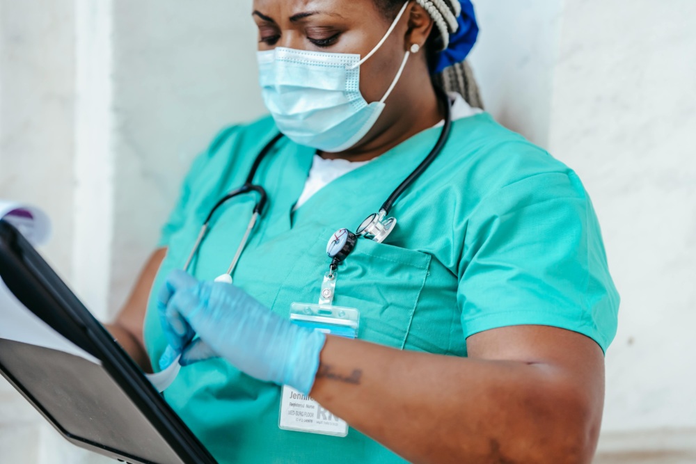 A healthcare worker in scrubs and a mask reviews a medical chart on a clipboard.